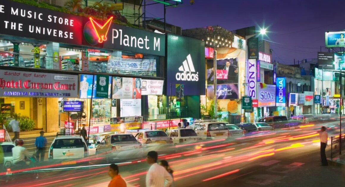 Night shopping scene on a busy commercial road in Bangalore, showing various brand stores and traffic