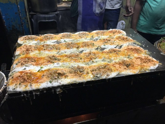 Specialty street food cooking on a hot griddle, showing long pieces of spiced egg or dosa batter topped with minced filling, common at night stalls in Chennai.